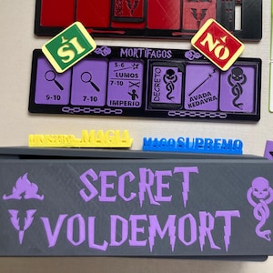 Puede incluir: Un tablero de juego con un diseño en negro y rojo. El tablero tiene un compartimento para guardar las piezas del juego. Las piezas del juego son rojas, negras y verdes. El juego se llama "Secret Voldemort".