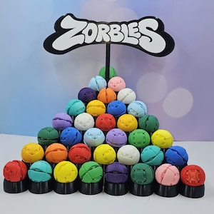 May include: A colorful, tiered display of spherical toys in various colors, including red, yellow, green, and blue. Each toy sits atop a black base. A sign at the top reads "ZORBIES" in white letters against a black background.