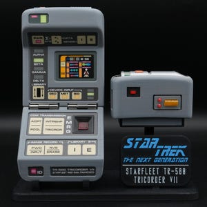 Op de afbeelding: Een grijze Star Trek TR-588 Tricorder VII prop replica. Het apparaat heeft een openklapbaar ontwerp met een scherm en diverse knoppen. Er is ook een kleiner, rechthoekig apparaat aanwezig. Een zwarte basis toont het Star Trek-logo en apparaatdetails.
