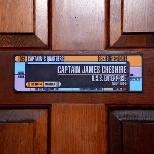 Op de afbeelding: Een rechthoekig bord voor de kwartieren van Captain James Cheshire op de U.S.S. Enterprise, gemonteerd op een houten deur. Het bord bevat de tekst "Captain's Quarters", "Deck 8 - Section 2" en "NCC-1701-D". Het bord heeft een zwarte achtergrond met kleurrijke randen.