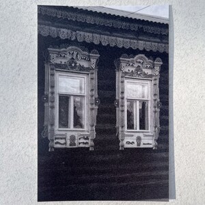 Puede incluir: Una fotografía en blanco y negro de un edificio de madera con dos ventanas ornamentadas. Las ventanas tienen tallas intrincadas alrededor y están enmarcadas por una cornisa de madera decorativa.
