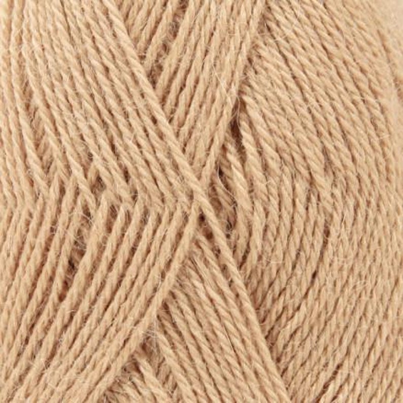 Puede incluir: Una vista de cerca de un ovillo de hilo de color tostado claro, que muestra la construcci&oacute;n de fibra trenzada y la textura suave y mate del hilo. Las hebras est&aacute;n enrolladas en un patr&oacute;n consistente, destacando el grosor uniforme y el color beige.
