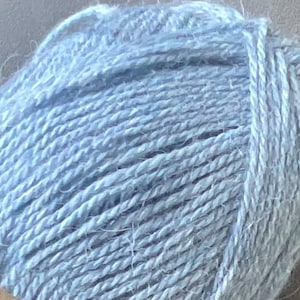 Puede incluir: Un ovillo de lana de color azul claro con una textura suave y esponjosa. El hilo est&aacute; enrollado en forma redonda, mostrando claramente las hebras retorcidas de la fibra. Una peque&ntilde;a parte de una etiqueta de papel tostado es visible abajo.