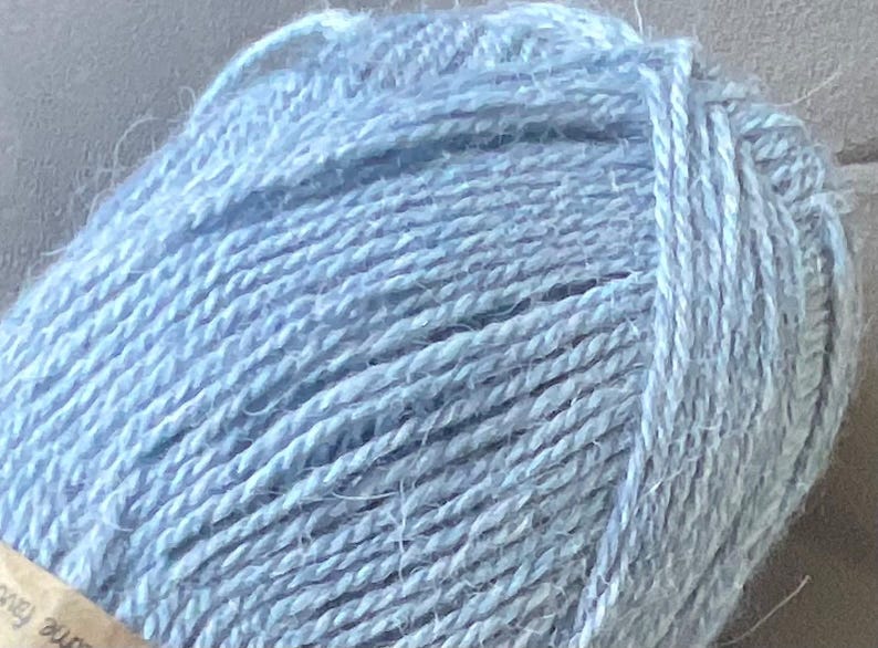 Puede incluir: Un ovillo de lana de color azul claro con una textura suave y esponjosa. El hilo est&aacute; enrollado en forma redonda, mostrando claramente las hebras retorcidas de la fibra. Una peque&ntilde;a parte de una etiqueta de papel tostado es visible abajo.