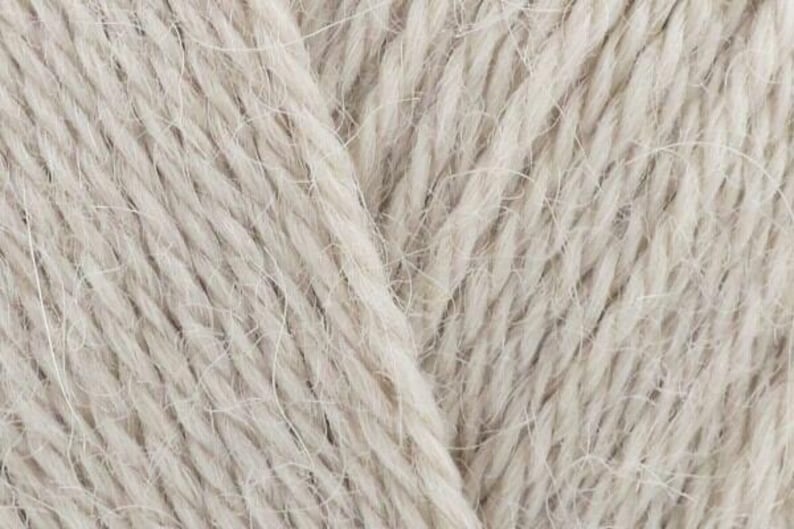 Puede incluir: Una vista de cerca de hilo de color beige claro con una construcci&oacute;n trenzada de m&uacute;ltiples cabos. Las fibras muestran una textura suave y ligeramente difusa con hebras individuales visibles tejidas juntas para crear un material textil neutro.
