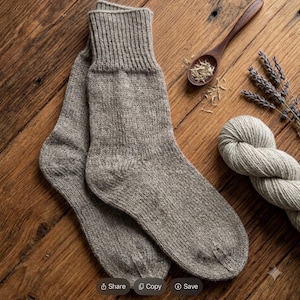 May include: A pair of light brown, knitted wool socks with a ribbed cuff. The socks are displayed alongside a wooden spoon containing dried herbs, a small bundle of dried lavender sprigs, and a skein of matching light brown yarn.