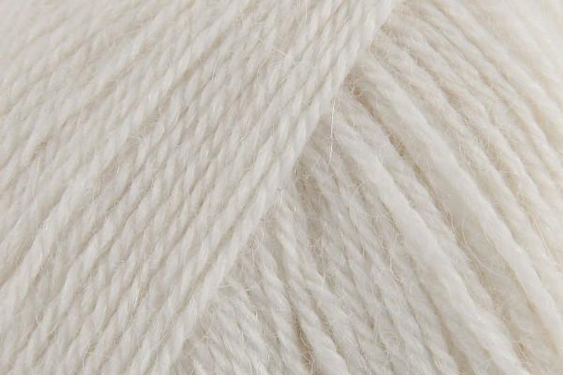 Puede incluir: Una vista de cerca de hilo color blanco roto con una construcci&oacute;n trenzada de m&uacute;ltiples cabos. Las fibras muestran una textura suave y ligeramente difusa con crestas diagonales visibles creadas por la torsi&oacute;n de los hilos.
