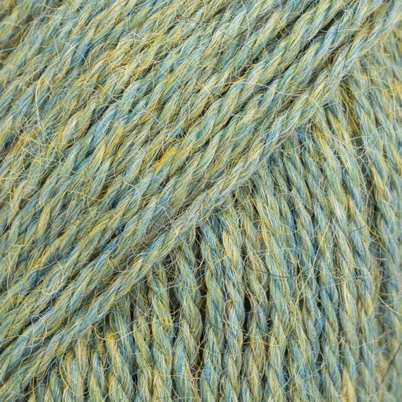 Puede incluir: Un hilo de varias hebras que presenta una mezcla de fibras de color azul suave y amarillo dorado retorcidas entre s&iacute;. La textura parece ligeramente difusa con una estructura de torsi&oacute;n visible, creando un efecto de color variado.