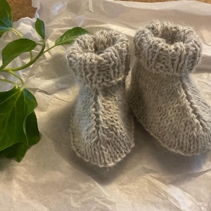 Puede incluir: Un par de patucos de bebé de punto gris claro. Los patucos están hechos de un hilo suave y texturizado y tienen un puño enrollado. Se presentan sobre papel blanco arrugado, con una planta de hojas verdes en el fondo.