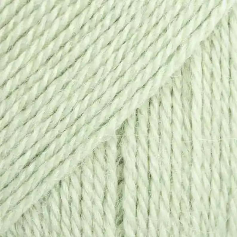 Puede incluir: Una vista de cerca de hilo de color verde menta claro con una construcci&oacute;n trenzada de m&uacute;ltiples capas. Las fibras exhiben una textura suave y ligeramente difusa con crestas diagonales visibles creadas por los hilos estrechamente enrollados.