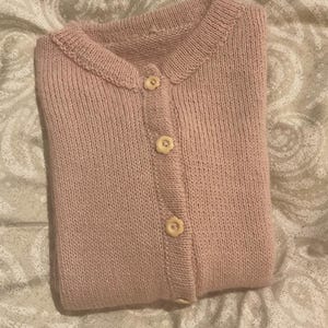 Puede incluir: Una rebeca de punto de color rosa claro con cuello redondo y cierre de botones en la parte delantera. La prenda est&aacute; confeccionada en punto liso y presenta tres botones de color crema con forma de peque&ntilde;as flores en el centro.