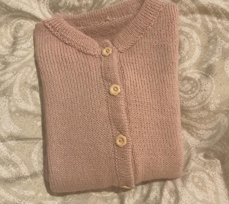 Puede incluir: Una rebeca de punto de color rosa claro con cuello redondo y cierre de botones en la parte delantera. La prenda est&aacute; confeccionada en punto liso y presenta tres botones de color crema con forma de peque&ntilde;as flores en el centro.