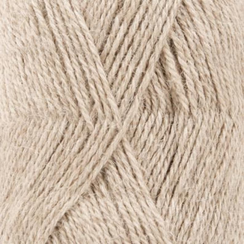 Puede incluir: Una vista de cerca de un ovillo de hilo de color beige claro, que muestra la estructura de fibra trenzada y la textura suave y difusa de los hilos individuales. El hilo est&aacute; enrollado creando un patr&oacute;n diagonal repetido en la superficie.
