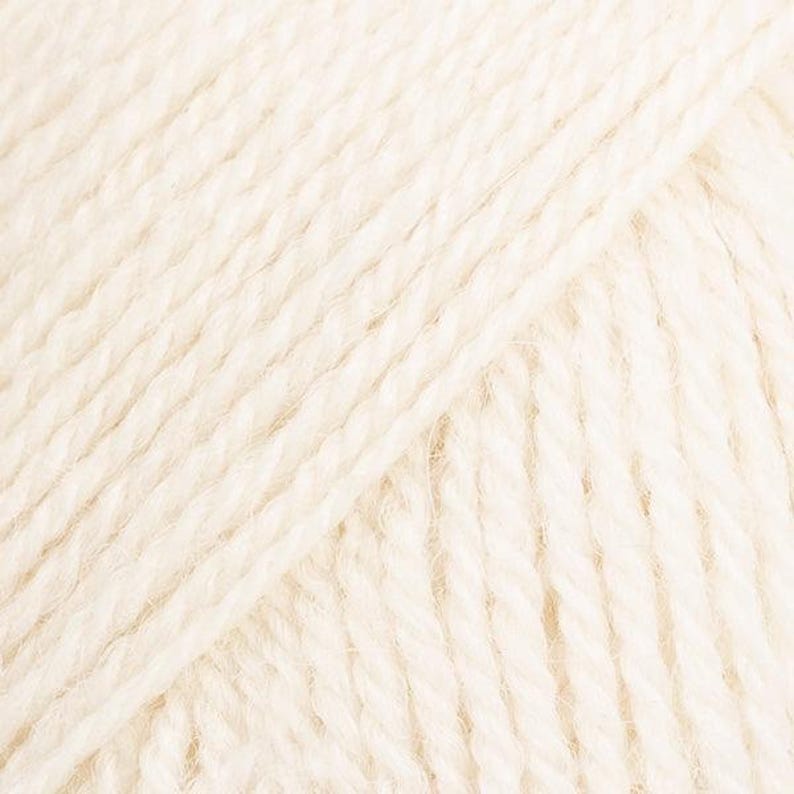 Puede incluir: Una vista de cerca de hilo color crema con una construcci&oacute;n trenzada de m&uacute;ltiples cabos. Los hilos individuales est&aacute;n fuertemente enrollados, creando una textura diagonal consistente en toda la superficie de la fibra, mostrando su tono natural.