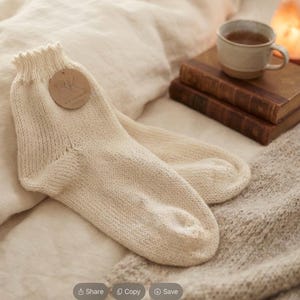 Könnte beinhalten: Ein Paar cremefarbene Strickstrümpfe mit geripptem Bündchen und einem runden braunen Papieranhänger, auf dem die Buchstaben RK aufgedruckt sind. Die Socken sind in glatt rechts gestrickt und haben eine weiche, strukturierte Oberfläche.