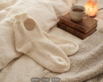 May include: A pair of cream-coloured knit socks featuring a ribbed cuff and a round brown paper tag with the letters RK printed on the surface. The socks are constructed with a visible stockinette stitch pattern and have a soft, textured appearance.