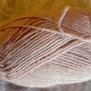 Puede incluir: Un ovillo de lana de color rosa p&aacute;lido con una construcci&oacute;n trenzada de m&uacute;ltiples cabos y una superficie ligeramente difusa y texturizada. Las fibras est&aacute;n enrolladas firmemente en forma ovalada, mostrando el color rubor uniforme.