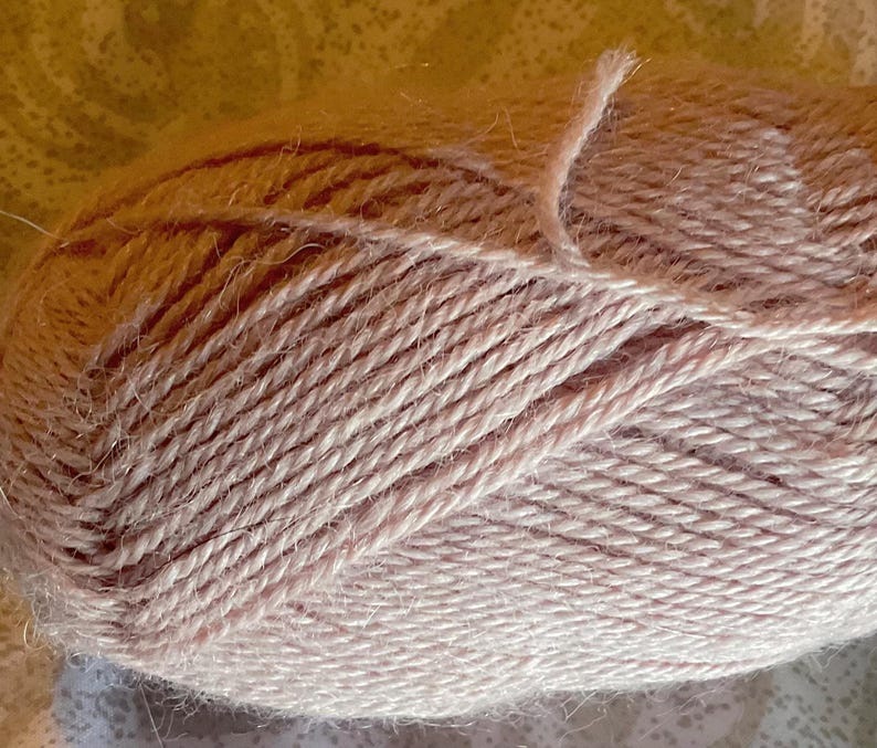 Puede incluir: Un ovillo de lana de color rosa p&aacute;lido con una construcci&oacute;n trenzada de m&uacute;ltiples cabos y una superficie ligeramente difusa y texturizada. Las fibras est&aacute;n enrolladas firmemente en forma ovalada, mostrando el color rubor uniforme.