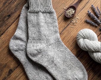 May include: A pair of light grey knit socks featuring a ribbed cuff and a textured, heathered appearance. The socks are displayed alongside a wooden spoon containing dried herbs, a sprig of dried lavender, and a ball of matching grey yarn.