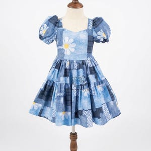 May include: A blue denim patchwork dress with white daisy accents. The dress features short, puffed sleeves and a tiered skirt. The bodice has a sweetheart neckline. The dress is displayed on a wooden mannequin.