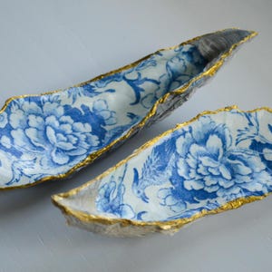 Op de afbeelding: Twee decoratieve, langwerpige schalen met een blauw-wit bloemmotief. De randen zijn afgewerkt met goud. De schalen zijn gemaakt van een textuurrijk materiaal en de binnenkant heeft een gedetailleerd bloemendesign.