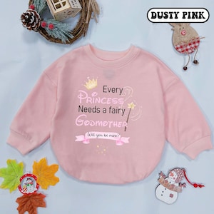 May include: A pink long-sleeved sweatshirt with a graphic print of a crown, a fairy wand, and the text "Every Princess Needs a Fairy Godmother. Will you be mine?"