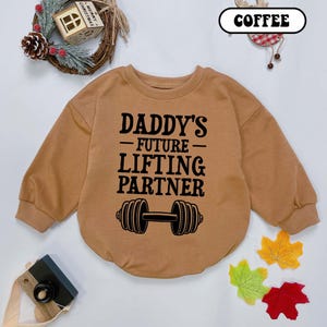 May include: A brown long-sleeved sweatshirt with the text "Daddy's -Future- Lifting Partner" and a black dumbbell graphic.