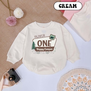 May include: Cream-colored baby onesie with long sleeves. The front features the name "Oliver" above a graphic that says "ONE Happy Camper" with a bear, tree, and wagon wheels. The word "CREAM" is in a white oval.