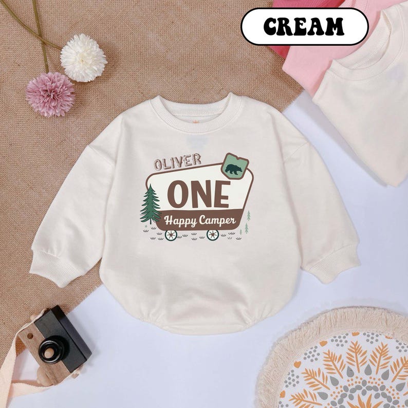 May include: Cream-colored baby onesie with long sleeves. The front features the name "Oliver" above a graphic that says "ONE Happy Camper" with a bear, tree, and wagon wheels. The word "CREAM" is in a white oval.
