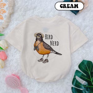 May include: A cream colored t-shirt with a cartoon illustration of a bird wearing a hat and binoculars. The text "Bird Nerd" is printed on the shirt.