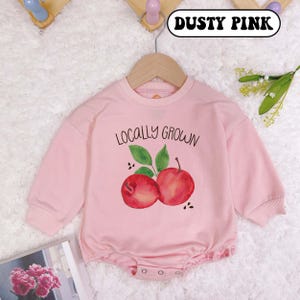 May include: A dusty pink long-sleeved baby onesie with a graphic of two red apples and the text "Locally Grown".