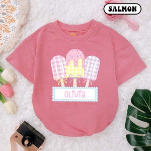 May include: A pink short-sleeved t-shirt with a graphic of three ice cream pops with a gingham pattern and sprinkles. The text "Olivia" is printed on a blue banner below the ice cream pops.