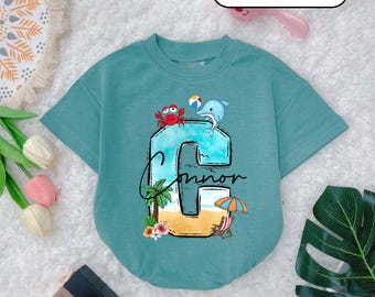 May include: A teal blue t-shirt with a summer beach theme graphic. The graphic features a large letter C with a beach scene, a crab, a dolphin, a palm tree, an umbrella, and the name "Connor".