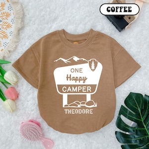 May include: A brown short-sleeved t-shirt with a white graphic of a sign that says "One Happy Camper Theodore".