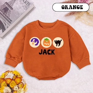 May include: Orange baby bodysuit with long sleeves. The front features three circular designs with a purple ghost, an orange jack-o'-lantern, and a black cat. The word "JACK" is printed below the designs. The word "ORANGE" is printed at the top.