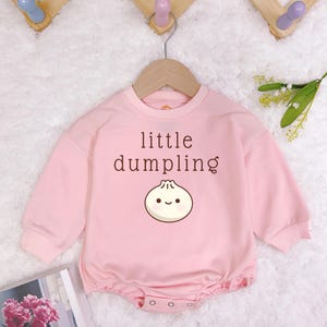 May include: A light pink baby romper with long sleeves and a round neckline. The romper features the text "little dumpling" above a cartoon dumpling illustration. The garment is hanging on a wooden hanger.