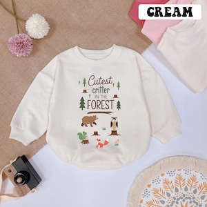 May include: A cream colored long-sleeved sweatshirt with a graphic of a bear, owl, and fox in a forest setting. The text on the sweatshirt reads "Cutest critter in the forest".