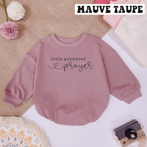 May include: Mauve taupe colored sweatshirt with long sleeves and a crew neck. The sweatshirt has the text "little answered prayer" with a heart symbol printed on the front.