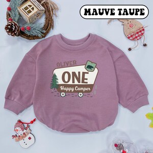 May include: Mauve taupe baby romper with long sleeves. The romper features the text "OLIVER ONE Happy Camper" with a bear and tree graphic. The romper is a solid mauve color.