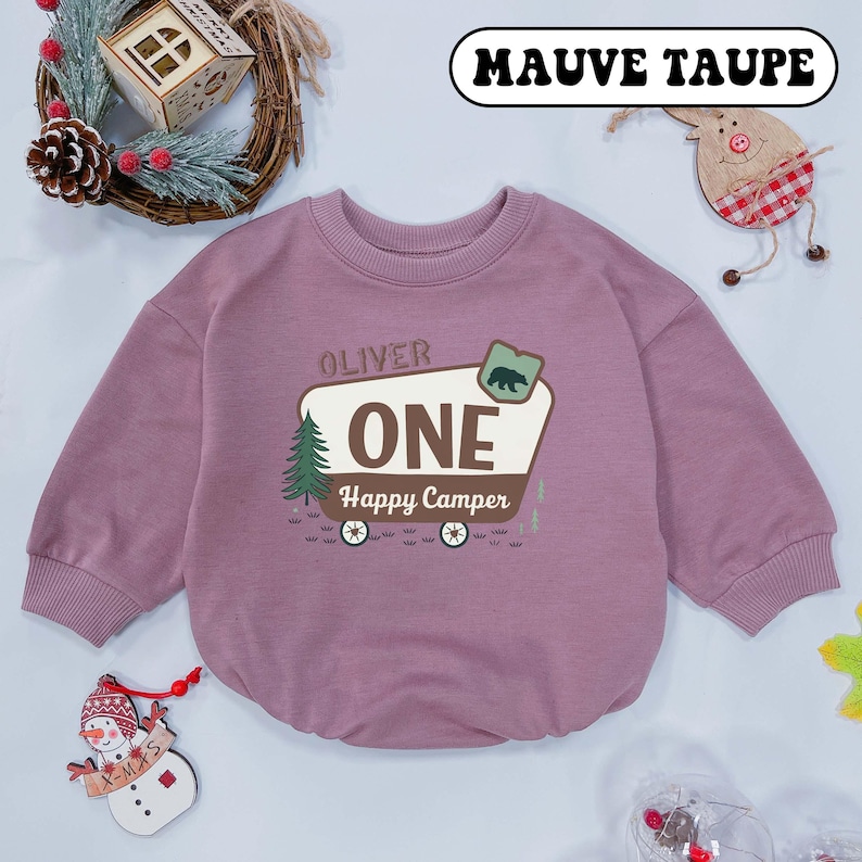 May include: Mauve taupe baby romper with long sleeves. The romper features the text "OLIVER ONE Happy Camper" with a bear and tree graphic. The romper is a solid mauve color.