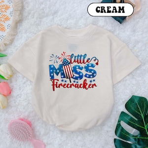 May include: Cream-colored baby bodysuit with the text "Little Miss Firecracker" in red, white, and blue. The design includes a firework and stars, perfect for celebrating Independence Day. The bodysuit has short sleeves and a round neckline.