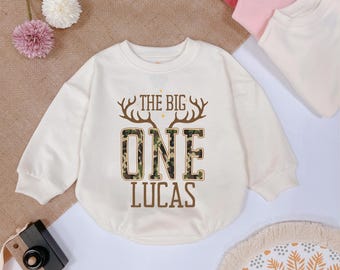 May include: A cream colored long-sleeved shirt with a camouflage pattern and the text "The Big One Lucas" printed on the front.