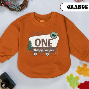 May include: Orange long-sleeved sweatshirt with a graphic that reads "OLIVER ONE Happy Camper." The graphic includes a bear, a pine tree, and a camper design. The word "ORANGE" is written in a white box at the top.