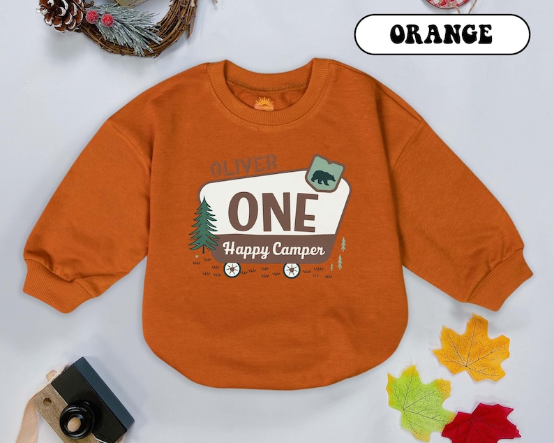 May include: Orange long-sleeved sweatshirt with a graphic that reads "OLIVER ONE Happy Camper." The graphic includes a bear, a pine tree, and a camper design. The word "ORANGE" is written in a white box at the top.