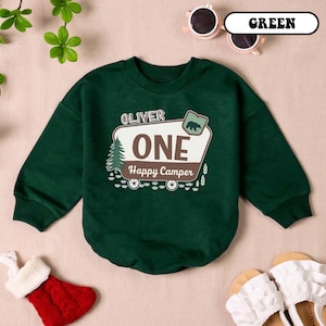 May include: A dark green sweatshirt with the text "OLIVER ONE Happy Camper" in a white and brown design. The design includes a bear, a pine tree, and wagon wheels. The sweatshirt is a crew neck style.