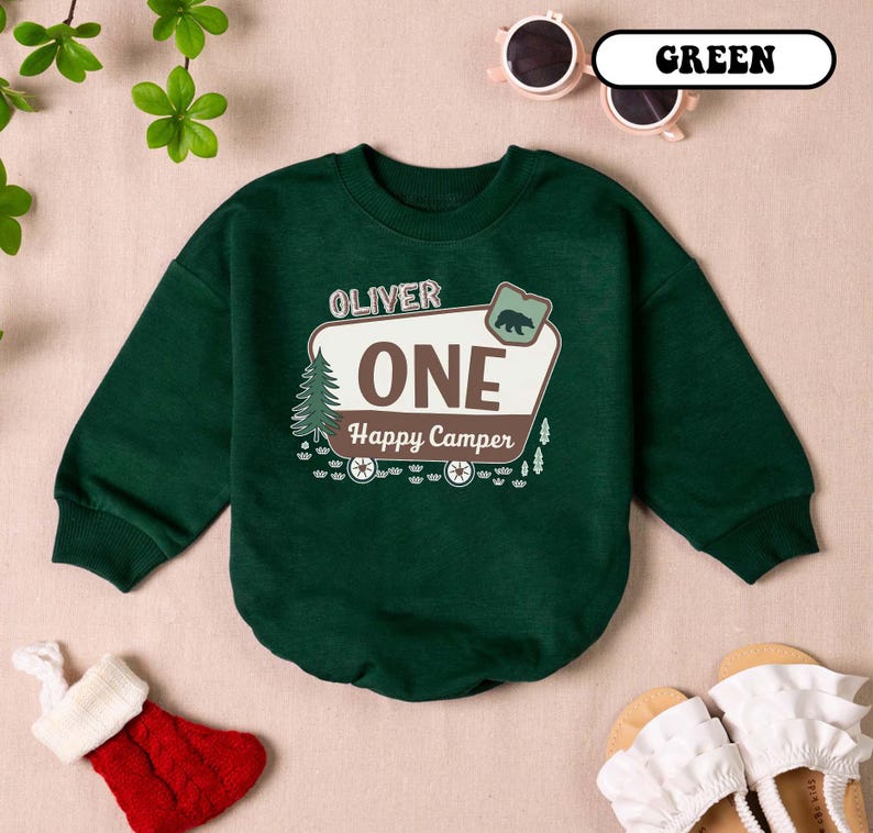 May include: A dark green sweatshirt with the text "OLIVER ONE Happy Camper" in a white and brown design. The design includes a bear, a pine tree, and wagon wheels. The sweatshirt is a crew neck style.