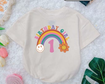 May include: A cream colored t-shirt with a rainbow, a smiley face, a flower, and the text "Birthday Girl 1" printed on it.