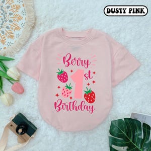 May include: A pink short-sleeved shirt with the text "Berry 1st Birthday" and a strawberry graphic. The shirt is dusty pink.