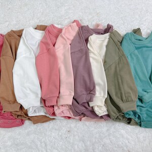 May include: A collection of baby rompers in various solid colors, including pink, brown, white, and teal. Each romper features long sleeves and a round neckline, arranged on a white, textured surface.