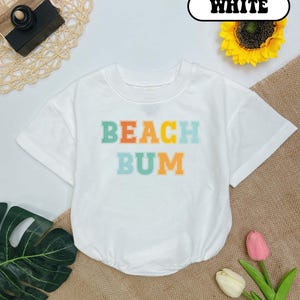 May include: White short-sleeved t-shirt with the text "BEACH BUM" in rainbow colors.
